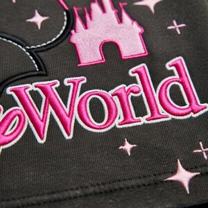 May include: Dark gray fabric with pink embroidered design elements. Features a pink castle, stars, and the word "World" in a stylized font. A pink balloon is also present. The design is likely for a themed apparel item.