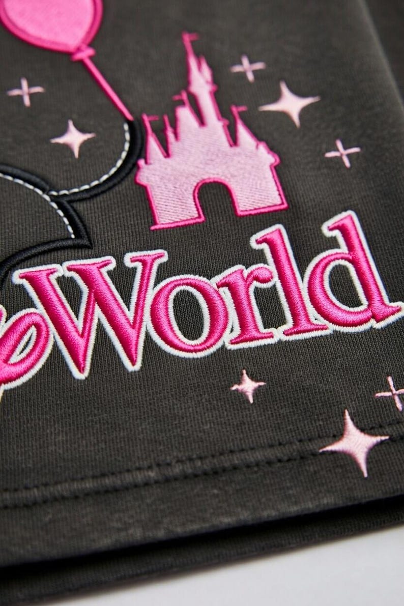 May include: Dark gray fabric with pink embroidered design elements. Features a pink castle, stars, and the word "World" in a stylized font. A pink balloon is also present. The design is likely for a themed apparel item.