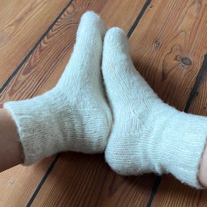 May include: A pair of off-white, hand-knitted ankle socks featuring a ribbed cuff design and a textured, fuzzy yarn finish. The socks are constructed with a visible heel gusset and a smooth knit pattern throughout the foot and ankle area.