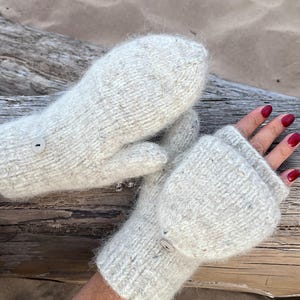 May include: A pair of off-white, chunky knit convertible mittens featuring a ribbed cuff and a small, round, light-coloured button on the back of each hand. The mittens have a fold-over flap design that allows the fingers to be exposed or covered.