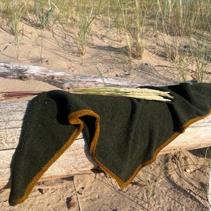 Pode incluir: Um xale triangular de malha em verde oliva escuro com uma borda contrastante em amarelo mostarda. O acessório têxtil está drapeado sobre um pedaço de madeira flutuante, com um pequeno feixe de talos de trigo seco por cima.