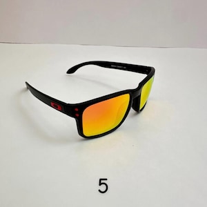 May include: Black sunglasses with orange mirrored lenses. The sunglasses have a rectangular frame with a red logo on the side. The lenses reflect a bright orange color. The number "5" is printed in black below the sunglasses.