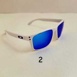 Pu&ograve; includere: Occhiali da sole bianchi con lenti blu a specchio. Le montature sono bianche con aste grigie. Il numero "2" &egrave; stampato in nero sotto gli occhiali da sole.