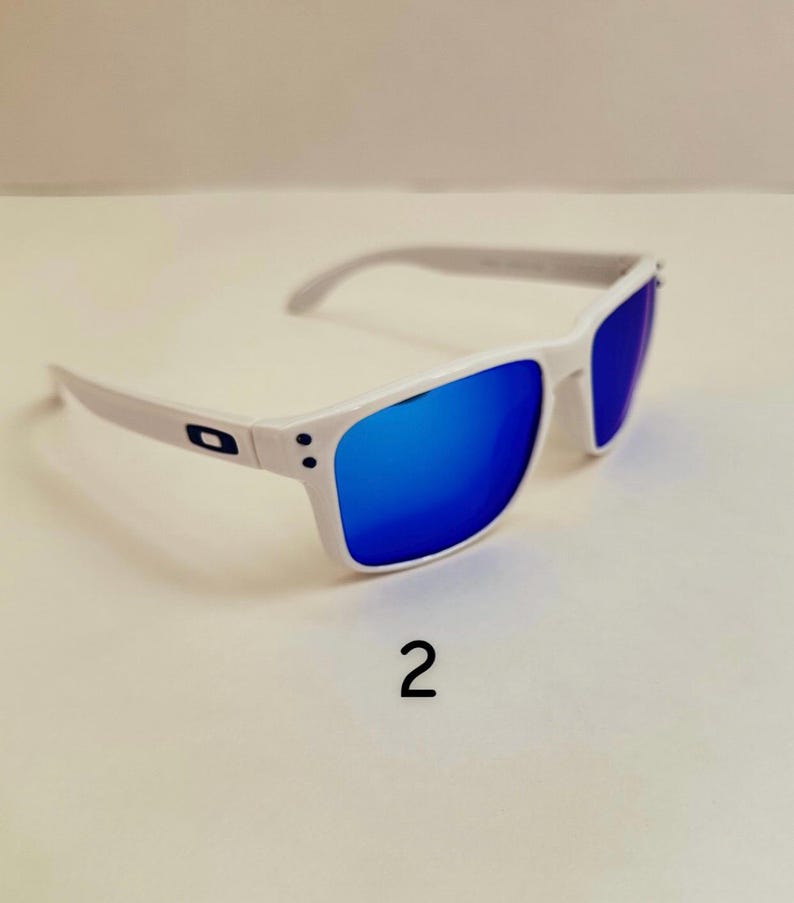 Pu&ograve; includere: Occhiali da sole bianchi con lenti blu a specchio. Le montature sono bianche con aste grigie. Il numero "2" &egrave; stampato in nero sotto gli occhiali da sole.