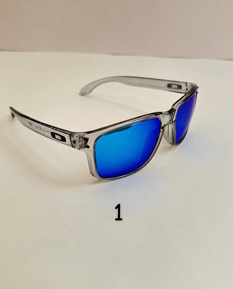 May include: Clear-framed sunglasses with blue mirrored lenses. The sunglasses have a rectangular shape and a transparent frame. The number "1" is printed in black below the sunglasses.