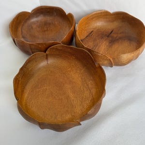 Può includere: Tre ciotole di legno con un design a petali. Le ciotole sono realizzate in legno marrone chiaro e hanno una finitura liscia e lucida. Le ciotole sono disposte su una superficie bianca.