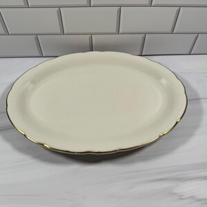 May include: An oval-shaped serving platter with a smooth, cream-colored surface and a scalloped edge. The rim of the platter is finished with a thin, metallic gold-colored trim that follows the curved contour of the plate's perimeter.