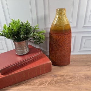 May include: A tall, cylindrical ceramic vase with a speckled orange and brown glaze, positioned next to two stacked hardcover books. The top book is dark red with the text "winemaking at home" and holds a small, metallic, ribbed planter.