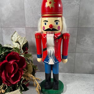May include: A wooden nutcracker figure wearing a red uniform with blue accents, a tall red hat with a gold star, and black boots. The figure has a white faux fur beard, a black mustache, and stands on a round green base.