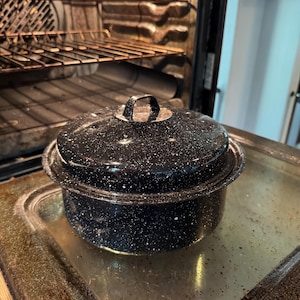 May include: A black enamel roasting pot with a lid, speckled with white dots, sits inside an oven. The pot has a handle on the lid and is on a metal surface. The oven's interior is visible.