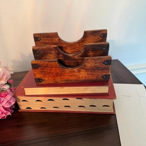 May include: A stack of three wooden letter holders with a curved top, resting on two red books. The letter holders are a warm brown color with dark metal accents. The books have gold-colored pages.