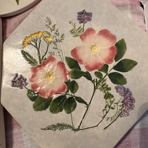 May include: An octagonal ceramic tile featuring a pressed floral design. The tile showcases pink and white roses, yellow wildflowers, and purple blossoms, with green leaves and stems. The flowers are arranged on a light-colored background.