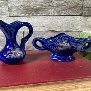 May include: A cobalt blue ceramic pitcher with a curved handle and a matching cobalt blue ceramic bowl featuring two side handles. Both pieces are decorated with a floral decal showing purple and white flowers with green leaves and gold trim.