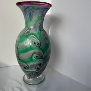 May include: A decorative glass vase with a flared top and a clear, rounded base. The vase features a swirling pattern of green, purple, and white colors. The rim of the vase is a deep magenta color.