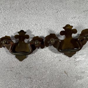 May include: Two matching brass drawer pulls featuring a decorative backplate with a central fleur-de-lis shape and curved, swinging handles. The metal has a dark, aged patina with visible highlights on the edges of the hardware components.