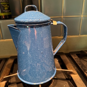 May include: A blue enamel coffee percolator with a white speckled pattern, featuring a matching lid with a small handle, a side handle, and a spout. The metal surface shows signs of wear along the edges of the lid, handle, and base.