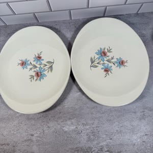 May include: Two oval-shaped off-white ceramic serving dishes featuring a central floral pattern. Each dish displays a cluster of light blue flowers with pink centers and green leaves, centered on the smooth, cream-colored surface of the bowls.