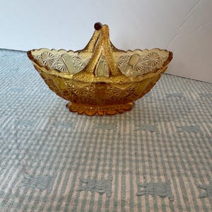 May include: Amber-colored glass basket with a handle and intricate starburst and geometric patterns. The basket has a scalloped rim and a decorative base. The handle is attached with a small, round, dark brown bead. A vintage decorative piece.