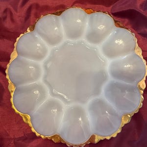 May include: A white, scalloped egg serving dish with a gold-colored rim. The dish has 12 individual egg compartments and a central serving area. The gold rim adds a touch of elegance to this vintage-style serving piece.