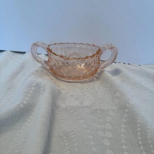 May include: A vintage pink glass sugar bowl with two handles. The bowl has a diamond pattern and a scalloped rim. The sugar bowl is made of clear glass and is sitting on a white cloth.