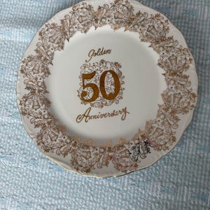 May include: A white ceramic plate with a gold rim and decorative gold filigree border. The plate features the words "Golden 50 Anniversary" in gold script, making it a commemorative piece.