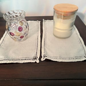 May include: Two rectangular off-white linen placemats with intricate openwork lace borders. One placemat holds a clear glass vase with colorful circular indentations, while the other holds a glass candle jar with a light-colored wax and a wooden lid.