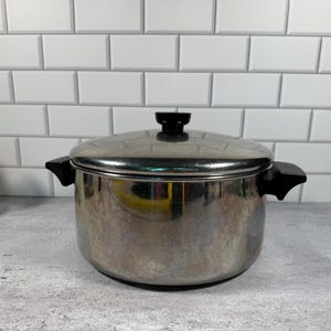May include: A stainless steel cooking pot with a polished finish, featuring two black side handles and a matching black knob on the lid. The pot has a cylindrical body and a flat lid that fits securely on top, showing reflective surfaces.
