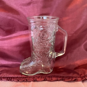 May include: Clear glass mug shaped like a cowboy boot. The mug has a handle and decorative embossed details. The glass is transparent, allowing the background to be visible. The mug is sitting on a red fabric.