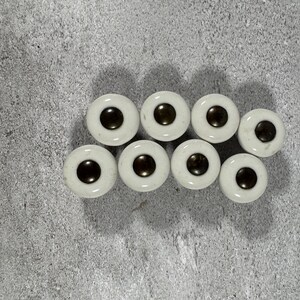 May include: A collection of eight circular buttons, each featuring a smooth, off-white ceramic base with a dark brown, slightly raised circular center. The buttons are arranged in two rows, with four buttons in each row, displaying a consistent design.