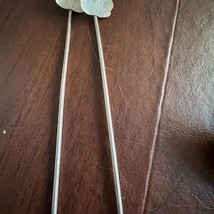 May include: Two silver-colored serving tongs with leaf-shaped ends. The tongs have long, slender handles and are resting on a brown surface. The leaf details are etched into the metal, adding texture to the design.