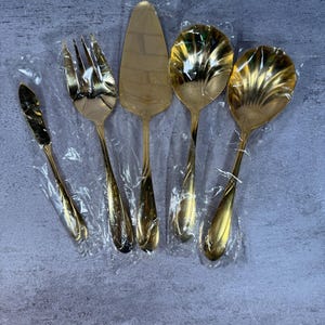 May include: A set of five gold-toned serving utensils, each individually wrapped in clear plastic. The collection includes a small knife, a four-tined fork, a triangular cake server, and two large serving spoons with scalloped bowl designs.