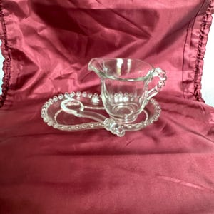 Puede incluir: Un juego de jarra de crema y platillo de vidrio transparente. La jarra de crema tiene un asa y un pico, y el platillo tiene un reposacucharas. Ambas piezas están decoradas con pequeños detalles en relieve en forma de cuentas. El juego se exhibe sobre una tela burdeos.