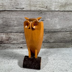 May include: A carved wooden owl figurine featuring a smooth, light brown body with two prominent circular eyes containing dark red, translucent centers. The owl stands on two thin wooden legs attached to a dark, textured, rectangular wooden base.