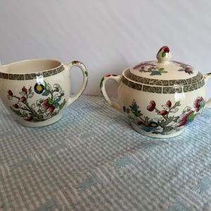 May include: Cream-colored ceramic creamer and covered sugar bowl set. Both pieces feature a floral design with red, blue, and green accents. The rims are bordered with a dark green geometric pattern. The sugar bowl has two handles and a knob on the lid.