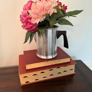 May include: A still life arrangement featuring a bouquet of pink and red flowers in a silver metal pitcher with a black handle. The pitcher rests on two stacked, maroon-colored books with yellowed pages. The arrangement sits on a dark wooden surface.