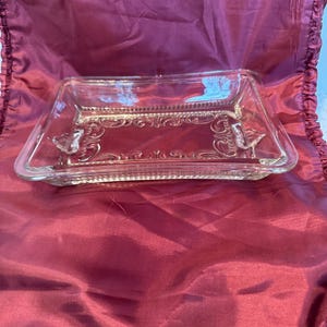 Può includere: Un piatto rettangolare di vetro trasparente con un motivo decorativo in rilievo al centro. Il piatto ha un bordo rialzato e piccoli accenti a farfalla su ciascuna estremità. È esposto su un tessuto bordeaux.