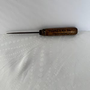 May include: An antique ice pick with a wooden handle. The handle is brown with the text "PEOPLE'S ICE CO. MANUFACTURED ICE" printed on it. The pick is made of metal and is pointed.
