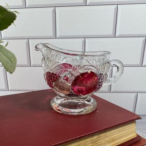 May include: A clear glass creamer pitcher featuring a raised, textured fruit motif with vibrant red and purple accents. The vessel has a curved handle, a pouring spout, and a pedestal base, showcasing detailed embossed leaves and fruit clusters.