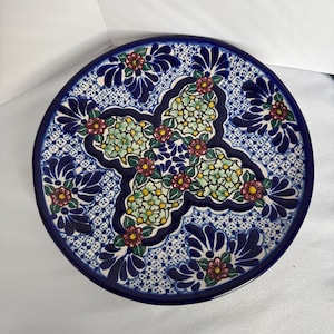 May include: A round, hand-painted ceramic plate with a blue and white floral design. The plate features a central four-petal floral motif with red, yellow, and green accents, surrounded by a detailed border of stylized flowers and patterns.
