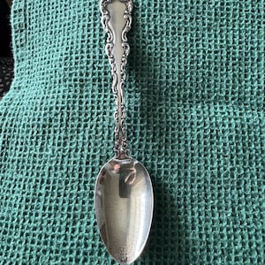 May include: A silver spoon with an ornate handle and a rounded bowl. The handle features a detailed, scrolling design. The spoon is set against a textured, teal green background, creating a contrast between the shiny metal and the matte fabric.