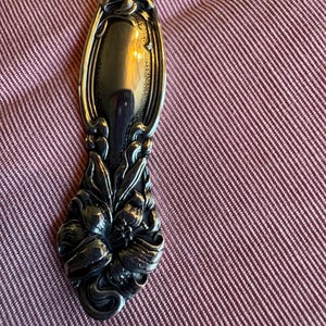 May include: A silver spoon with ornate floral detailing. The spoon's bowl is oval-shaped and reflects the surrounding environment. The handle features a detailed floral design, adding a touch of elegance. The spoon is set against a red background.