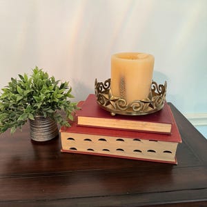 May include: A decorative candle holder with a large, beige candle sits atop two red books. The candle holder is made of gold-colored metal and has an ornate design. A small potted plant is to the left.