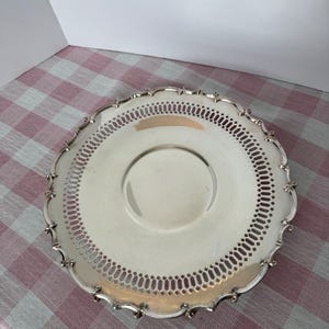 Puede incluir: Una bandeja redonda plateada con un diseño calado decorativo alrededor del borde. La bandeja tiene un borde festoneado y un centro ligeramente elevado. La bandeja está sobre una superficie a cuadros rojos y blancos.