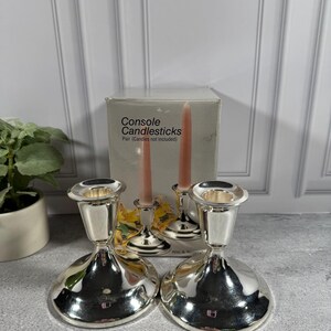 May include: A pair of silver-toned metal console candlesticks with a wide, circular base and a tapered stem. Behind the candlesticks is a gray cardboard box labeled Console Candlesticks Pair (Candles not included) with an image of the product.