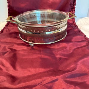 Puede incluir: Un plato de servir de vidrio transparente con base y borde de metal plateado. El borde presenta un patrón decorativo de pequeños rombos. El plato tiene asas de madera y se apoya en pequeños pies de metal. Fondo de tela roja oscura.