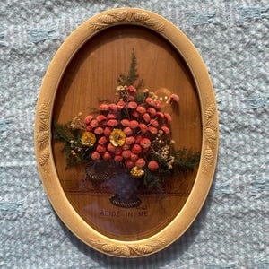 May include: Oval-shaped framed artwork featuring a floral arrangement in a basket. The arrangement includes red, yellow, and green dried flowers. The frame is a light beige color with a decorative pinecone design. The text "ABIDE IN ME" is visible.