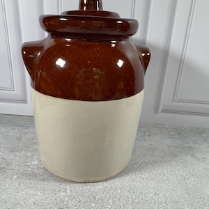 May include: A two-toned ceramic crock featuring a glossy dark brown glaze on the upper portion and a smooth cream-colored finish on the lower body. The vessel includes two side handles and a matching removable lid with a central knob handle.