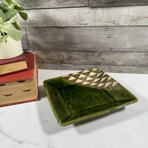 May include: A square ceramic dish featuring a glossy, deep green glaze on the majority of its surface, contrasted by a corner section decorated with a repeating pattern of cream and dark brown triangles, creating a distinct geometric visual effect.