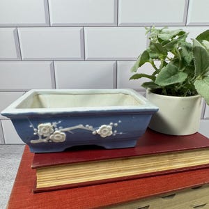 May include: A rectangular ceramic bonsai pot with a light blue glazed exterior featuring a raised white floral branch relief. The interior is white, and the pot has a wide rim and a flat base. It is a functional container for small plants.