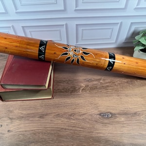 May include: A cylindrical wooden rainstick with a polished light brown finish and dark speckles. It features two black bands with white geometric patterns and a central black sun illustration with rays. The instrument rests on two stacked books.